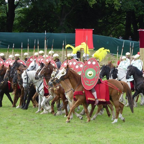 Reenactors form a line of Roman cavalry on a grassy field, armed with spears, oval-shaped shields, shining helms and full-body armour.