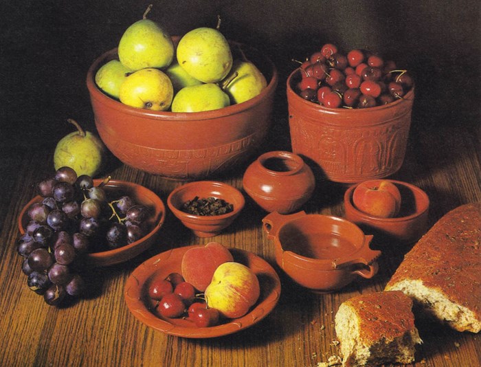 Reddish-brown vessels on a table overflowing with pears, grapes, bread, raisins and peaches