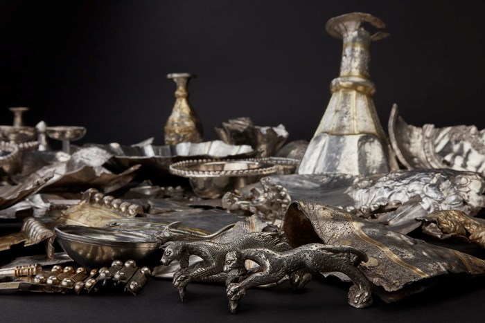A vast array of silver objects laid on a black surface. Two big cat figures are in the foreground, with many vessels, cups and shards scattered around them.