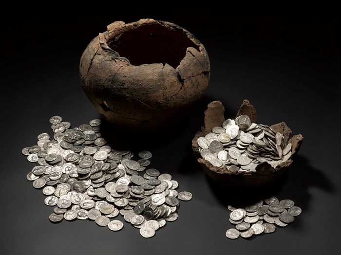A broken pot surrounded by several piles of silver coins set against a dark background