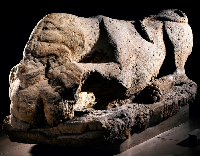 Side view of a large stone sculpture of a monstrous-looking lioness lying down while feasting on a human figure