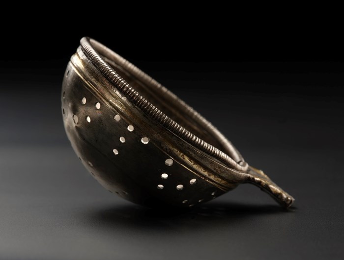 A shallow dished strainer with holes in its side and rope-like metal rim leaning on its side against a dark background.