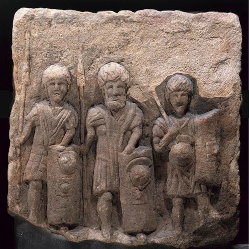 Stone block depicting three boldly carved figures of soldiers standing side by side and looking straight ahead, armed with spears and shields. They feel like friends.