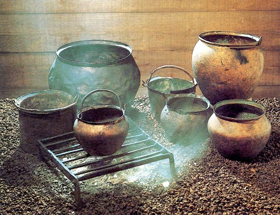 A bronze cooking vessel rests atop a grill with a small, smoky fire underneath it. Other vessels are positioned around the fire inside a cosy wooden building.