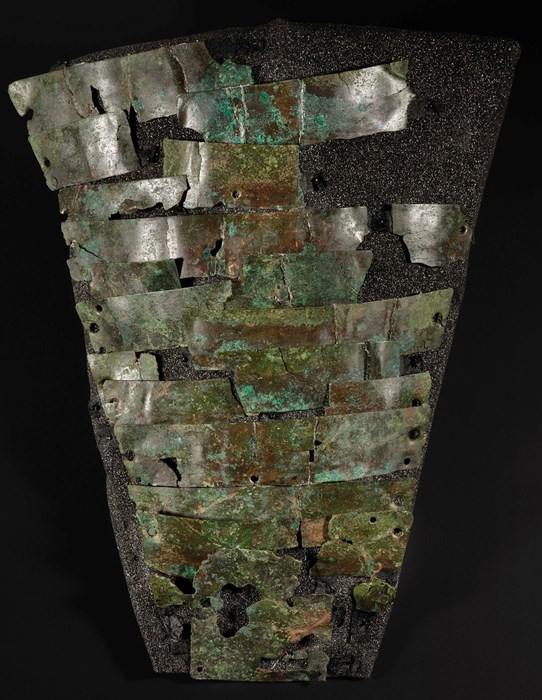 Rusted, speckled-green rectangular metal plates fastened together forming a protective layer over protective foam shaped like an arm