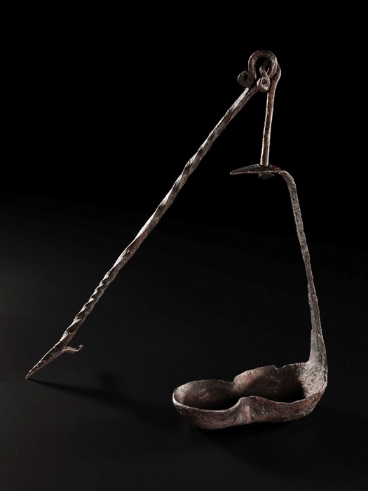 Rusted, brown-black hanging lamp. Basin forms a figure-eight shape, with thin stem leading up to a ring from which a spear-like appendage juts out