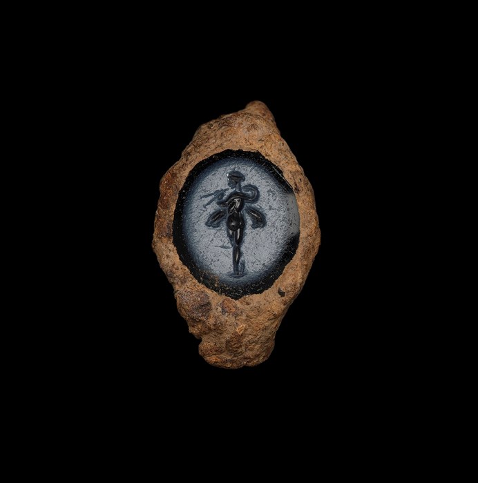 Small blue-grey gemstone set within a badly rusted, light brown ring. Gemstone has a nude figure holding tongs surrounded by swirling, wing-like patterns