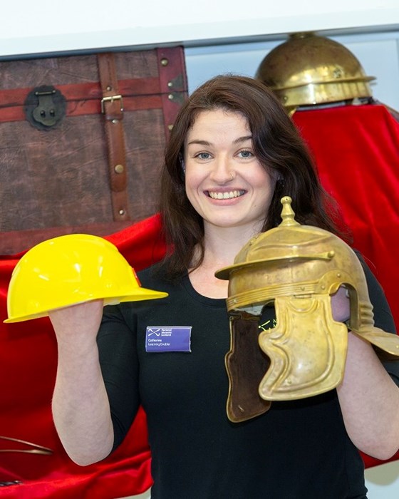 Smiling woman holding a modern yellow hard hat on the left and a replica Roman legionary helmet on the right