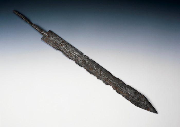 Rusted and damaged but largely intact sword blade against a pale background. Width is constant until a sudden and sharp triangular point at the end.