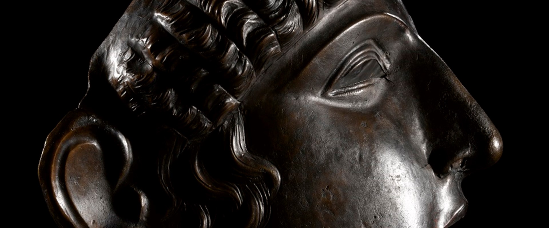 Profile view of bronze mask placed on a table. Light glints off its right cheek, and it bears a dignified expression.