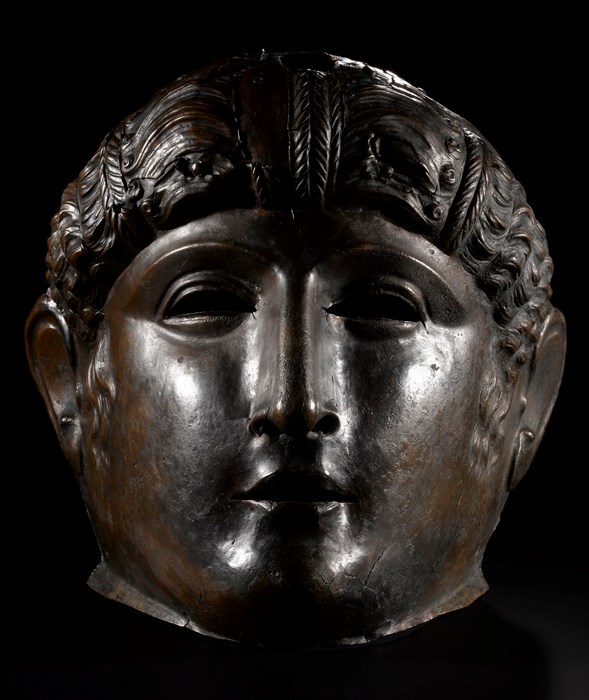 Darkened bronze mask staring straight ahead. Smooth, puffy features with a small mouth and nose, wavy hair and broad cheeks.