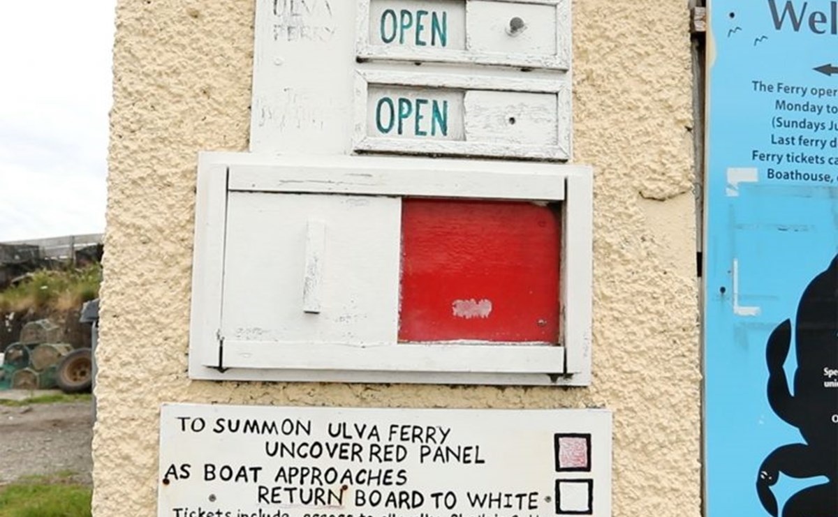 A wooden board reading 'to summon Ulva Ferry uncover red panel. As boat approaches, return board to white. Tickets include access to all walks, sheila's cottage and Ferry crossing. Tickets (returns_ Adult £6.00, Child £3.00, Cycle £0.50.' There is a sliding panel that is surrently revealing a red square. 