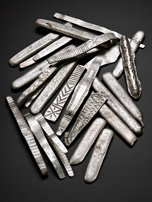 Pile of over 20 silver flattened arm-rings and finger-shaped ingots laid out on a black surface. Some are plain, others have chevron, cross and zipper-like patterns.