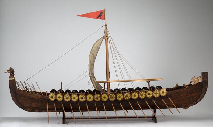 Scale model of a Norse longship. Yellow shields are slung over the ship's rim. Oars extend out at regular intervals, and a red flag flies from the lone, central mast.