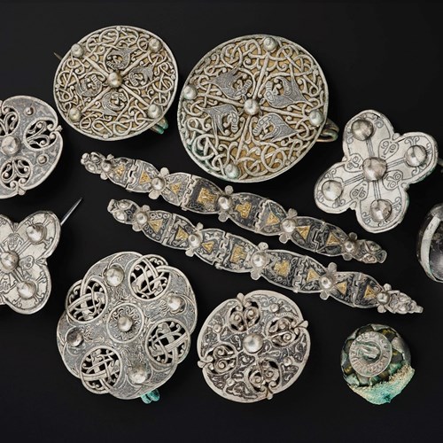 Cluster of silver objects on a black surface viewed from above. Two decorated metal strips in the centre with 9 brooches, mostly round, surrounding them.