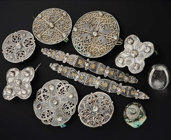 Cluster of silver objects on a black surface viewed from above. Two decorated metal strips in the centre with 9 brooches, mostly round, surrounding them.
