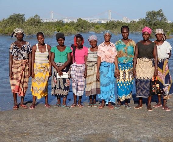 Group of nine women standing on the shore of a river on a sunny day. All facing forward, colourfully dressed. 