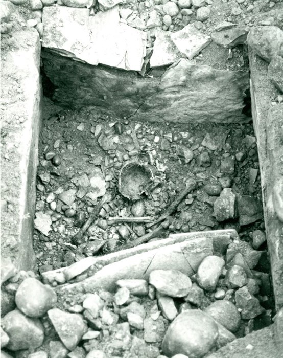 Black and white picture of a shallow rectangular pit lined with stone slabs. Inside is stone rubble, several human bones and pottery fragments.