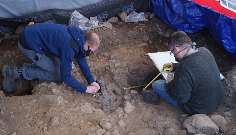 Two men excavate an archaeological pit, one on hands and knees and the other seated at the edge. They look focused and are flanked by tarps.