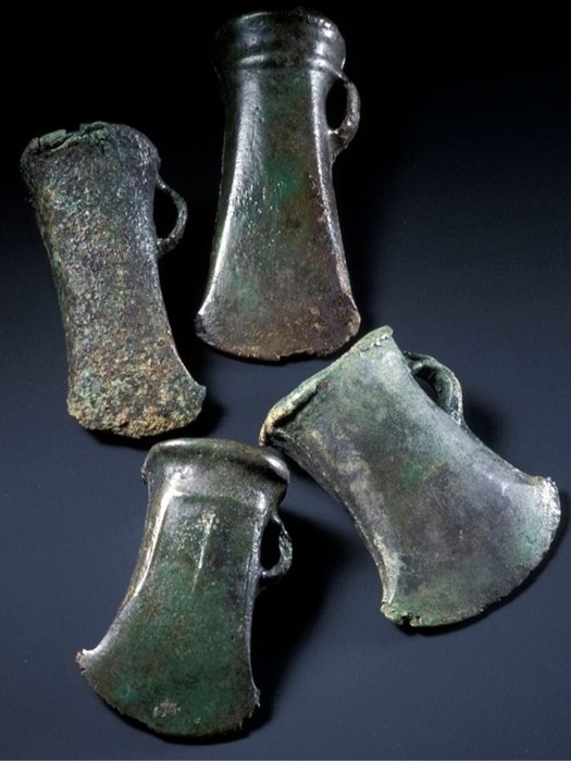 Four axe heads on a dark blue surface. Tinted green, their blades face toward the viewer. One is roughly textured, the rest are smooth.