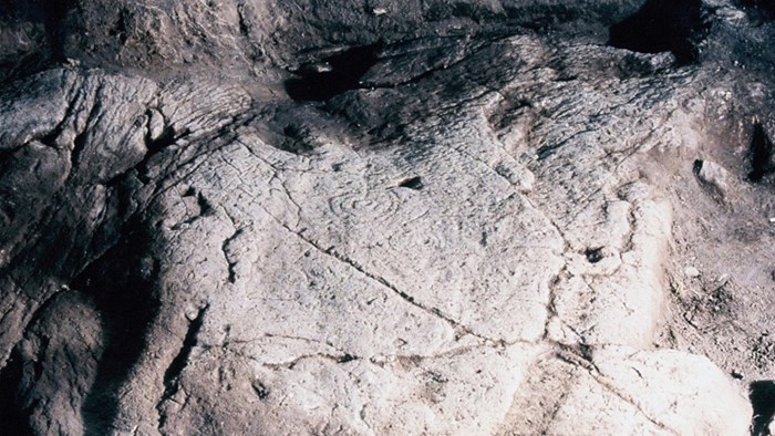 A light grey rock face illuminated at night time reveals cup-and-ring rock carvings on its uneven surface.