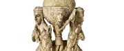 Tall off-yellow ivory sculpture. Two figures on horseback flack two orbs, on atop the other. A small human figure stand between the orbs, another crowns the sculpture.