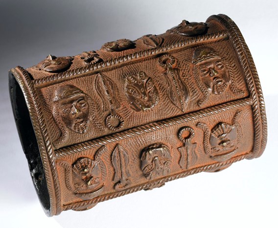 Brown-orange bronze armlet, shaped like a tube, decorated with bold symbols in relief.