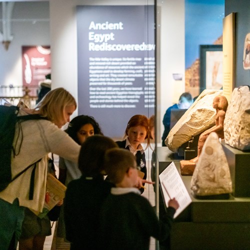A teacher gathers a group of primary pupils around one of the cases in the Ancient Egypt Gallery. 