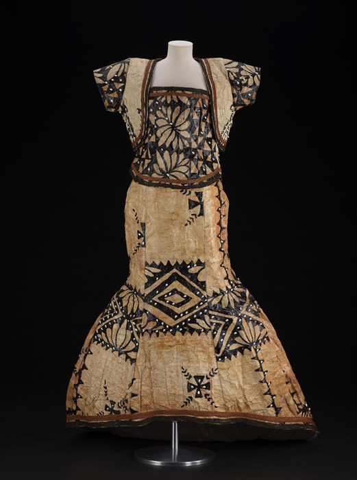 Pale brownish-yellow dress with black designs of flowers and geometric shapes. No sleeves. Mounted on a mannequin against a black background.
