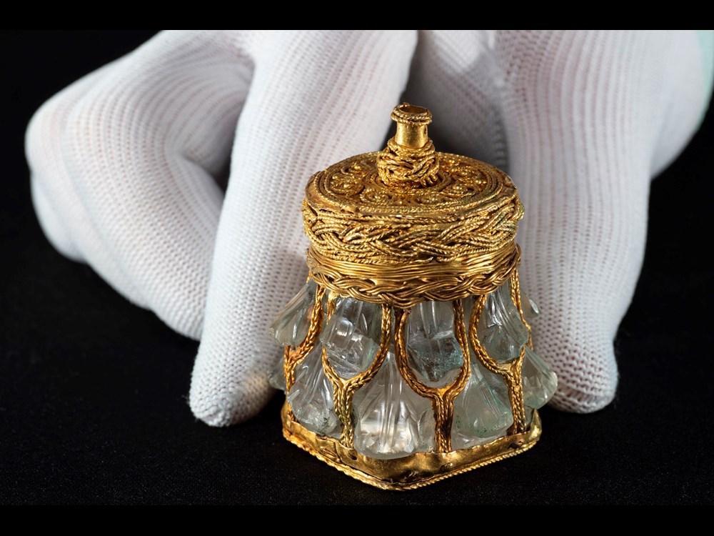 Light green crystal jar with gold base, lid and mesh-like covering against a black background with hand for scale. Jar is no taller than a finger.