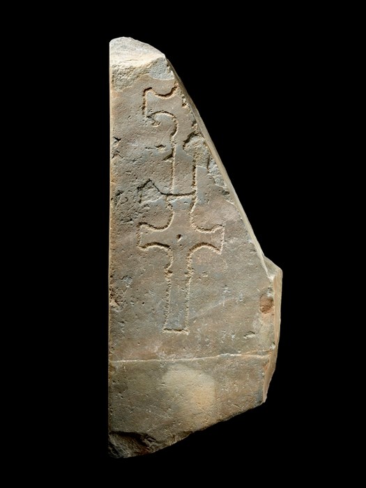 Roughly triangular slab of yellow-grey rock against a black background. Two simple crosses, one atop the other, are carved on it.