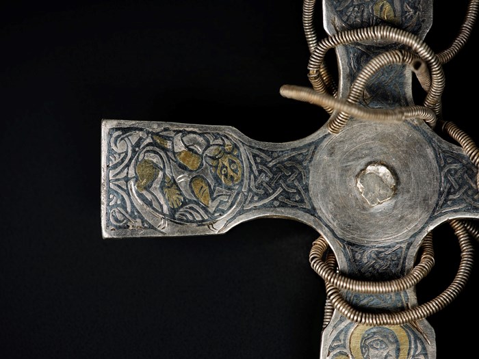 A silver cross cropped to focus on its centre and left arm. Plain black background. A bear-like creature is carved in black and gold on the left arm and a cord wraps around the centre of the cross.