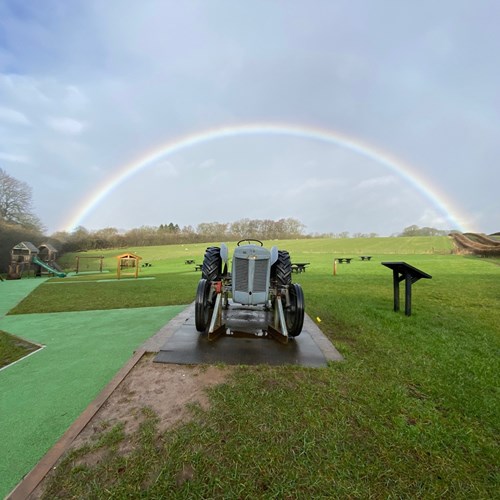A model tractor sitting in a playpark with a rainbow in the sky in the background.