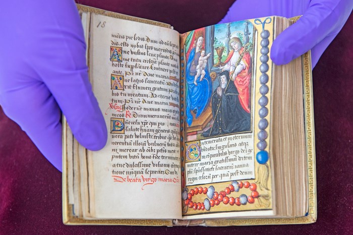 Pair of purple gloved hands holds a very old book open. Pages are filled with colourful Latin script and a vivid biblical scene on the right.