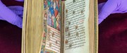 Very old open book with medieval script and a colourful Biblical scene, held open by two purple-gloved hands.