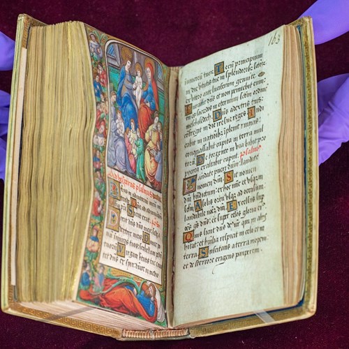 Very old open book with medieval script and a colourful Biblical scene, held open by two purple-gloved hands.
