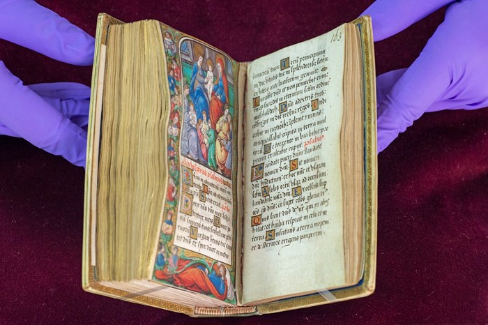 Very old open book, with beautiful Latin script on the right page and a colourful Biblical scene on the left, held open by two purple-gloved hands.