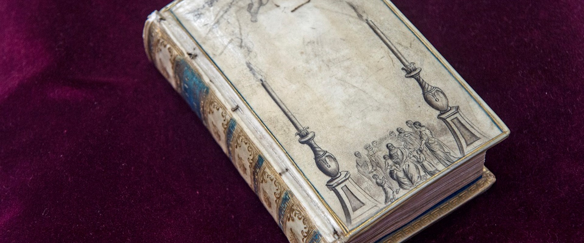 Very old book with a colourful spine on a velvety red surface. Faded off-white cover decorated with a cherub and group of people between two columns.