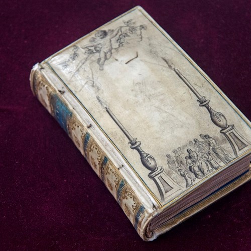 Very old book with a colourful spine on a velvety red surface. Faded off-white cover decorated with a cherub and group of people between two columns.
