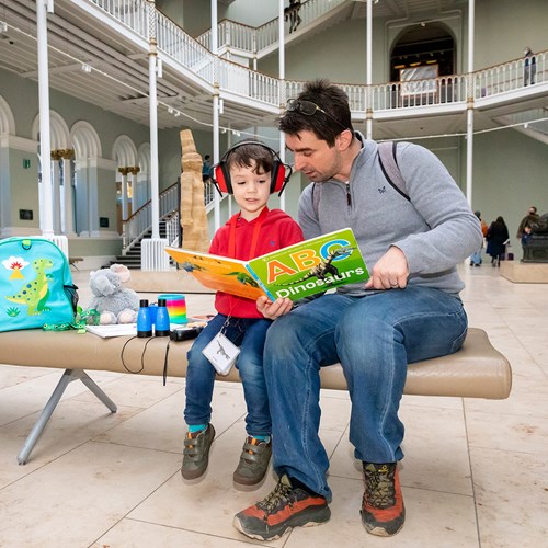 A man and a boy sit on a bench in the grand gallery looking at a large dinosaur picture book. The boy is wearing ear protectors.
