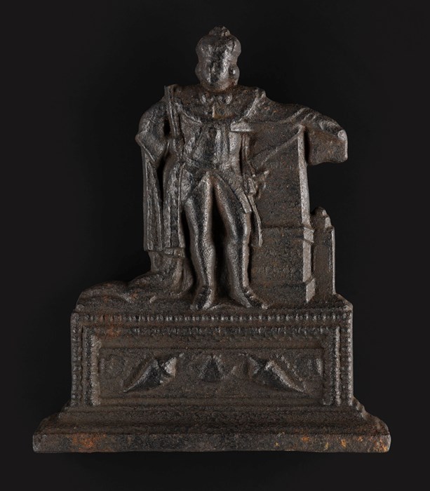Metal fireside ornament coloured black-brown. Figure wearing a crown and fancy robes stands atop a large plinth, leaning on a column.
