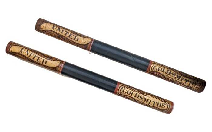 Two shot wooden batons side by side, black in centre with wood and gold ends. Left ends read 'United', right ends read 'Goldsmiths'.