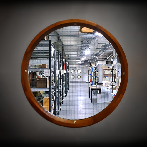A small circular window, like those you would see in the hull of a ship, gives a glimpse into a museum storage area filled with racks containing boxes and folders.