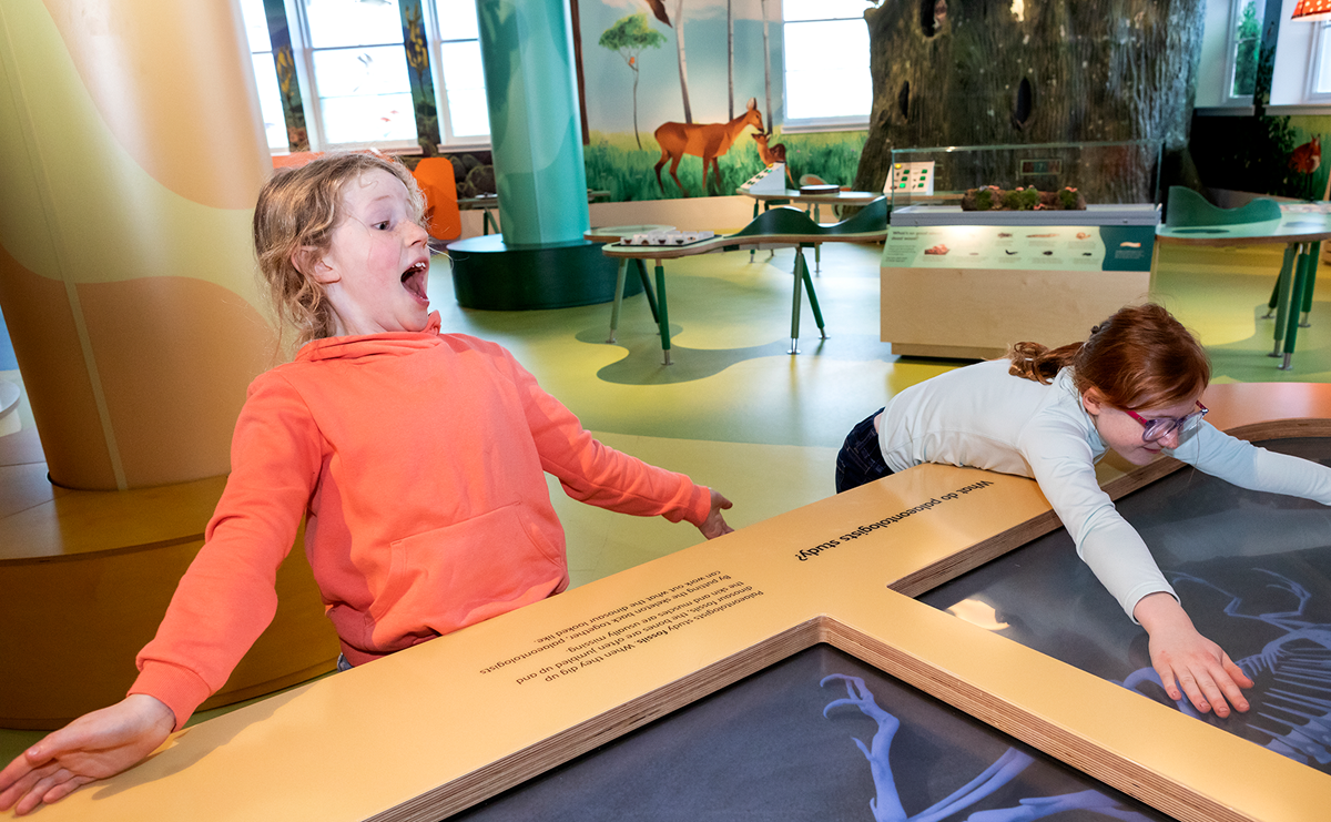 Two children in a gallery, one leans back with a look of surprise, the other rests on the display in front.