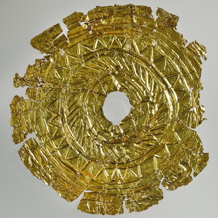 Gold foil disc, thin as paper laid out flat. Ziz-zag designs arranged in a spiral decorate the surface.