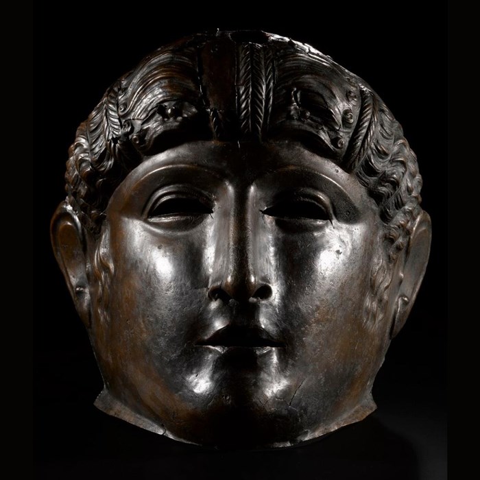 Metal face mask, dark grey-brown, of a young Roman man with strong nose bridge, curly hair and slightly open mouth.