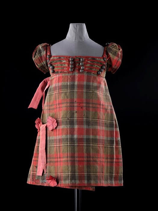 Young boy's tartan dress with pink bows, pink and green tartan, and cross-stitching over chest. Mounted on mannequin against black background.