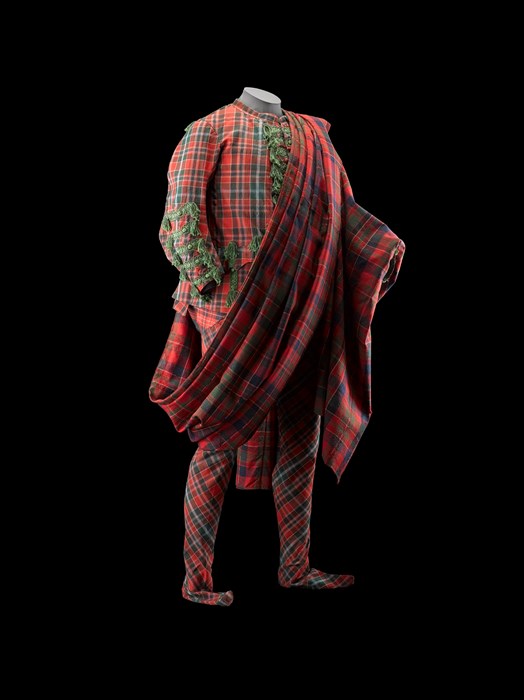 Full tartan suit from neck to toe, standing against black background. Dense red and green checkered pattern with large swoop of cloth across the torso.