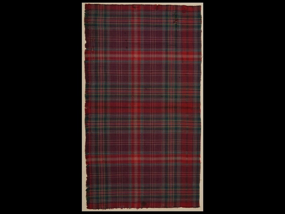 Tall rectangular swatch of burgundy, dark blue, red and light green tartan fabric against a black background.