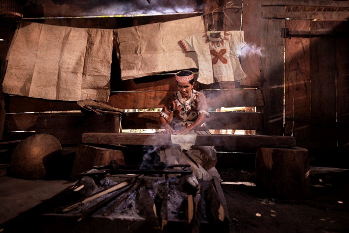 A woman in a brown barkcloth garment works steaming fabrics inside a wooden structure, with beige barkcloth sheets hanging on the walls .